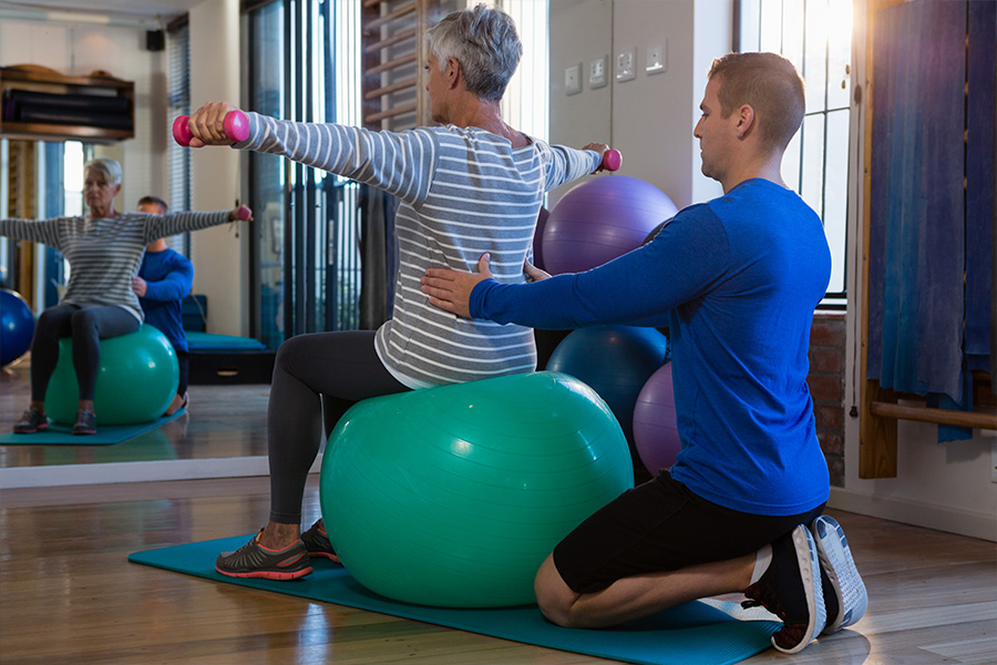 Physiotherapeut assistiert älterer Dame, die auf einem Medizinball sitzt und mit Hanteln krankengymnastische Übungen macht
