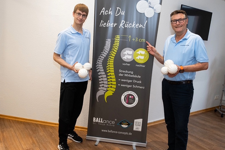 Physiotherapeut Mike Kiekseier und Mitarbeiter Pascal stehen in der Praxis Physio-Point in Hamburg vor einem Aufsteller zum BALLance Konzept von Tanja Kühne unter dem Slogan Ach Du lieber Rücken! Sie haben die dazu passenden Trainingsbälle in der Hand.